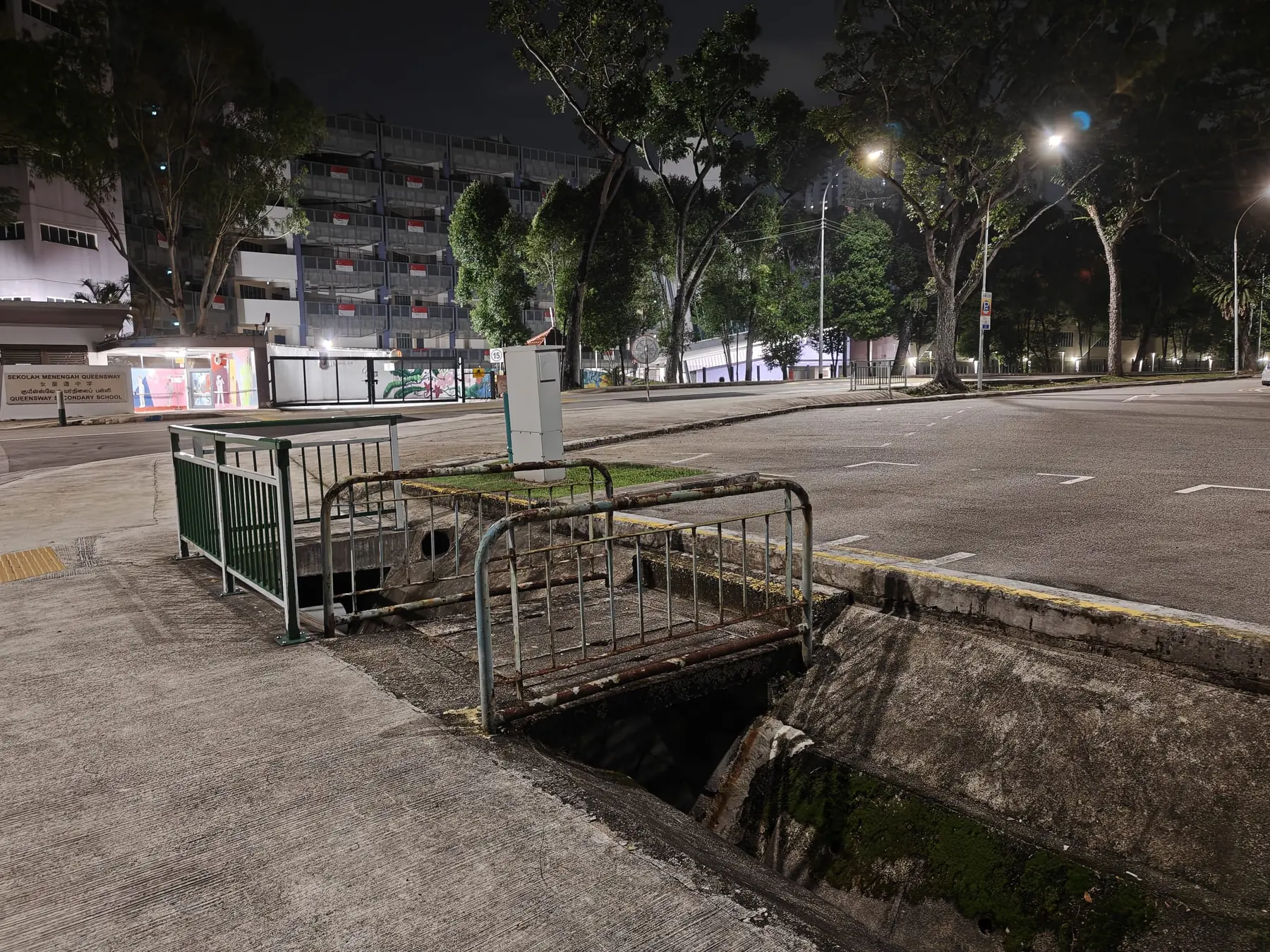 A concrete access crossing over a monsoon drain at night in Singapore, railed on both sides, leading into a car park that already has entrances. Queensway Secondary School and an HDB block hung with flags stand across the empty road.