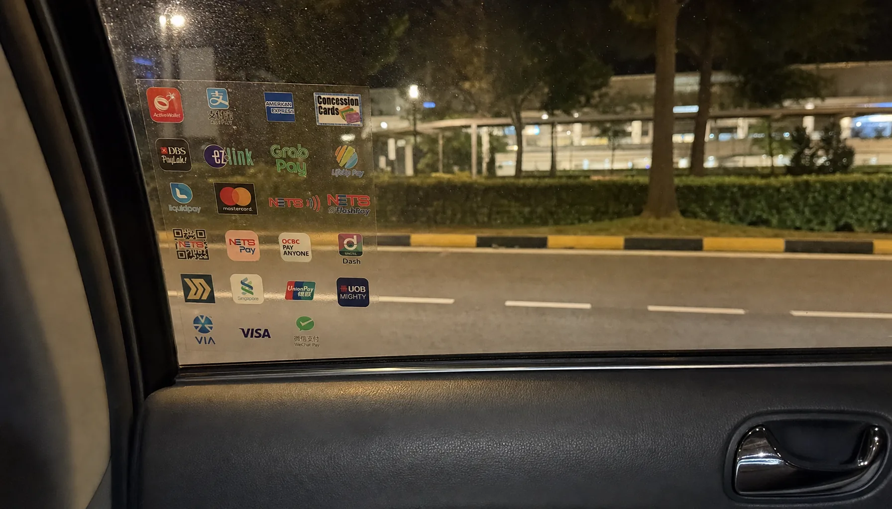 The inside of a Singapore taxi at night. A sticker on the window is crowded with two dozen payment logos - NETS, EZ-Link, PayLah, GrabPay, AliPay, Visa, UnionPay, WeChat Pay and more - with an empty, streetlit road outside.